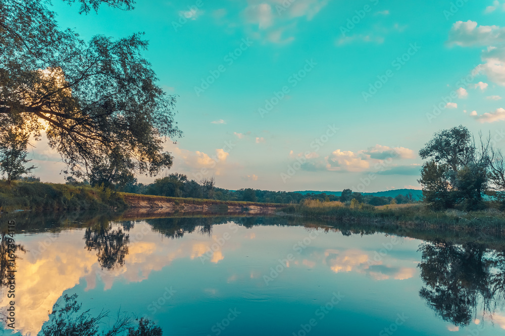 Calm beautiful scenic landscape with blue river, green trees and reflecting in water with cloudy sky. Magical sunset over the river in rural terrain. Natural, wild landscape. Calm beautiful scenic landscape with blue river, green trees and reflecting in water with cloudy sky. Magical sunset over the river in rural terrain. Natural, wild landscape.