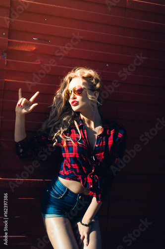 portrait of a girl close-up of a beautiful young sexy blonde in a red plaid shirt and shorts denim in sunglasses smiling and posing on the Sunset hipster
