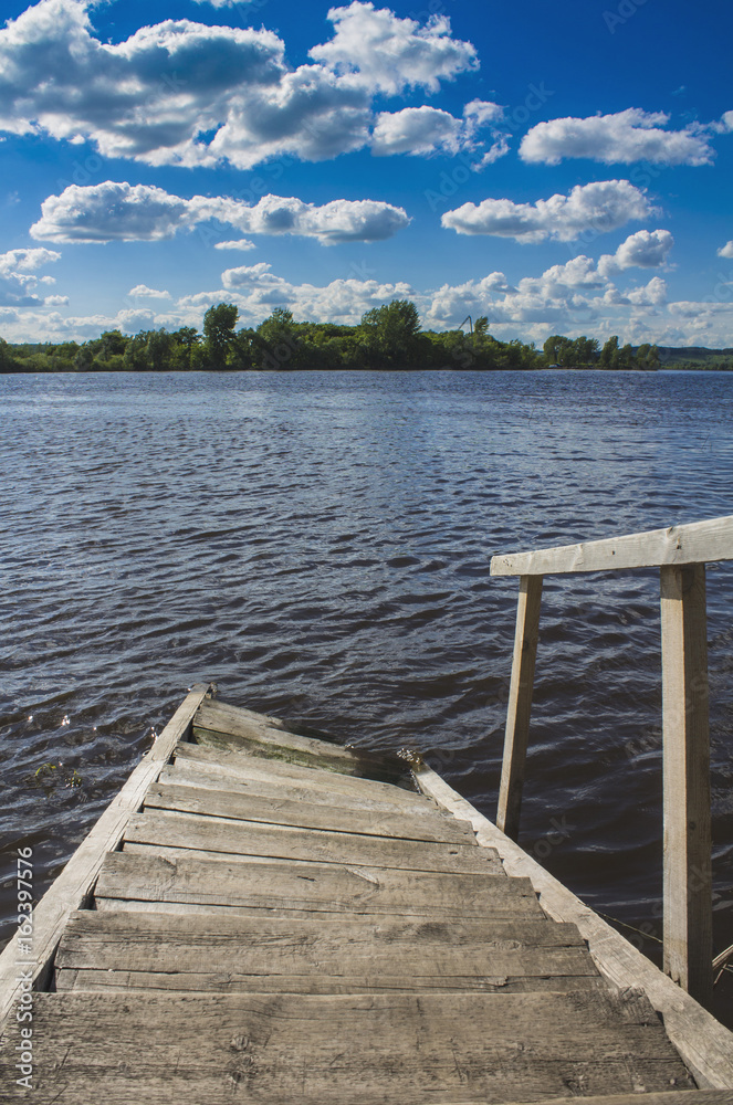 Obraz premium Wooden steps for boarding a boat go into the water under a blue sky with white clouds.