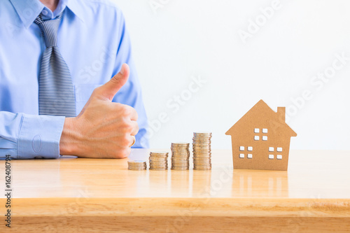 Business concept, saving planning for the future with model of the house and stack of coins on wooden table over blurred businessman with thumb up background