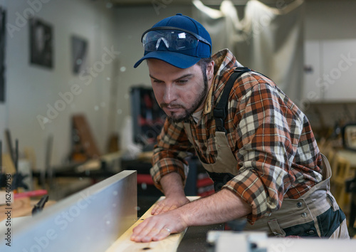 Young attractive white male in coverall and plaid shirt, working on woodworking machine in the carpenter workshop