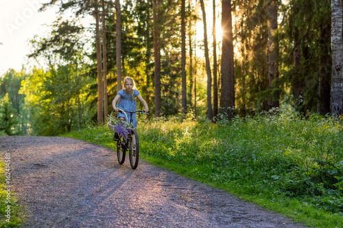Girl riding a bicycle in the woods with a basket of flowers, evening light