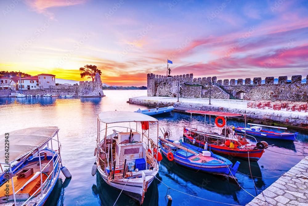 Old Greek port Nafpaktos with ancient harbor walls an background and fishing boats at foreground. Epic lilac colored sunrise sky over sea scenery. Nafpaktos is famous travel destination in Greece.