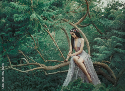 Fantasy girl in a fairy garden. A young princess in a purple dress sits on a green tree branch. Creative colors artistic processing. Fantasy woman image elf queen, forest nymph. Sexy gown costume