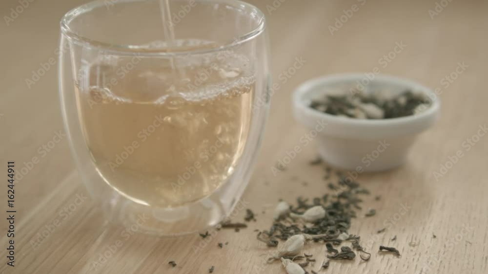 Green Jasmine Herbal Tea being poured into a glass or cup on wood bench top, showing dried loose leaf tea leaves and Jasmine flowers. High quality Pro Res 422 HQ footage. Green Jasmine Herbal Tea being poured into a glass or cup on wood bench top, showing dried loose leaf tea leaves and Jasmine flowers. High quality Pro Res 422 HQ footage.
