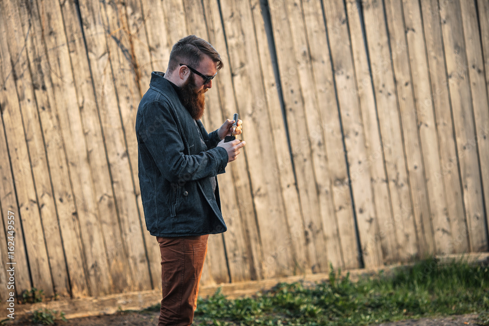 Fototapeta premium Vape man. E-liquid filling process. Outdoor portrait of a young brutal white guy with large beard vaping an electronic cigarette opposite the old wooden fence in the village.