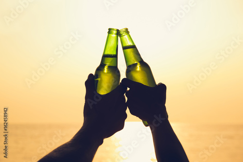 Couple hands holding beer bottles, clanging at the beach in summer sunset