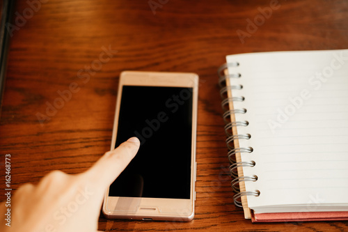 hand of woman touch on black screen of smartphone. blank notebook paper putting on beside. both putting on wooden table. image for technology,business,education,mobile,body part concept