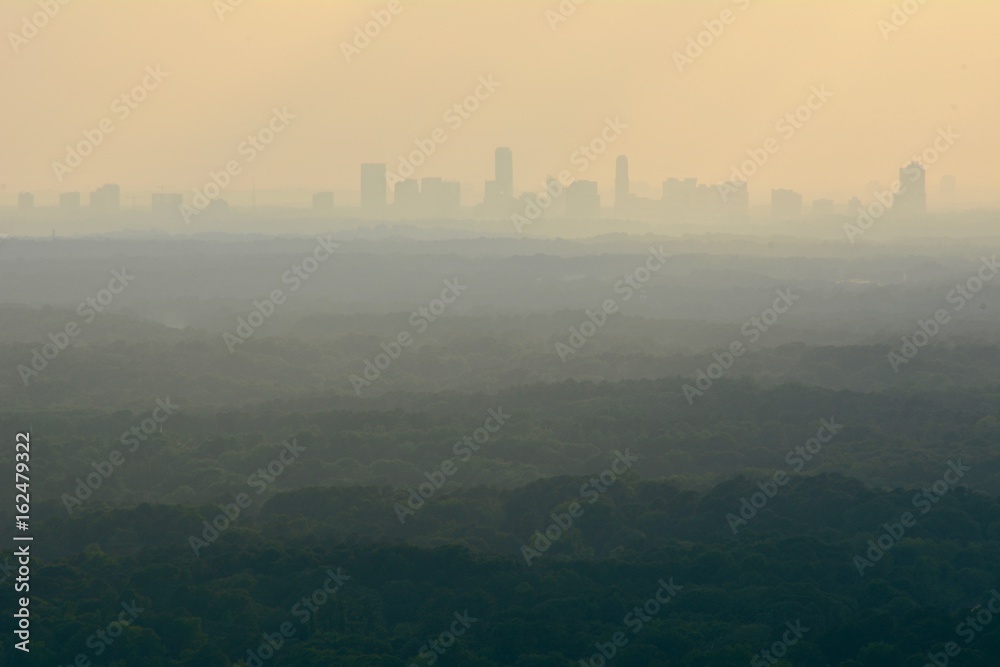 Cityscape or view of a city downtown from a distance with many layers of forests in the middle and orange sky showing sun rays shot at dusk from a mountain