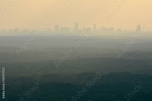 Cityscape or view of a city downtown from a distance with many layers of forests in the middle and orange sky showing sun rays shot at dusk from a mountain