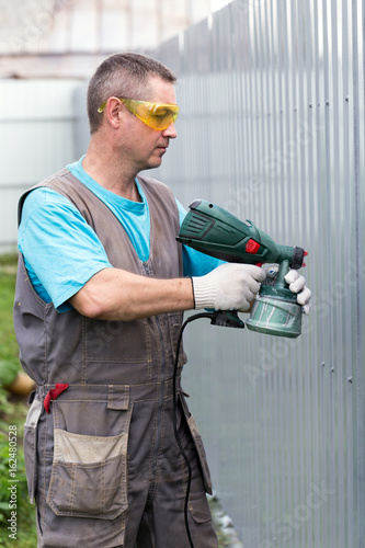Man in overalls and protective glasses with spray gun in hand. Painting of metal fence