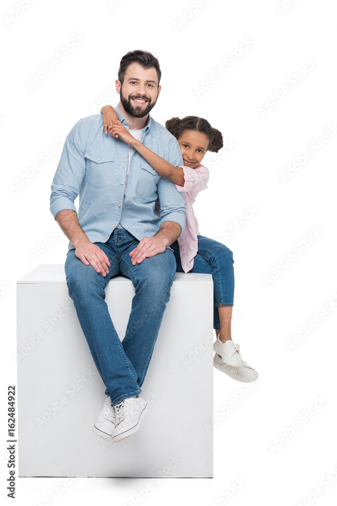 daughter hugging smiling father while sitting on cube together isolated on white daughter hugging smiling father while sitting on cube together isolated on white
