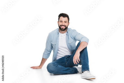 Handsome young bearded man sitting and smiling at camera isolated on white
