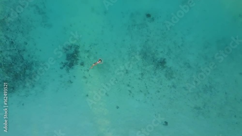 Aerial camera is ascending over an attractive woman in a bikini floating in crystal clear sea at atoll tropical island resort. Pretty sexy girl swimming in Indian ocean photo