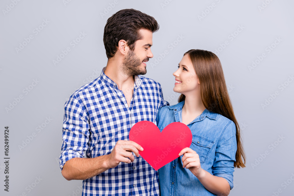 Love story. Trust and feelings, emotions and joy. Happy young lovely couple in love is posing, wearing casual shirts, holding big red paper heart and smiling on the pure background Love story. Trust and feelings, emotions and joy. Happy young lovely couple in love is posing, wearing casual shirts, holding big red paper heart and smiling on the pure background