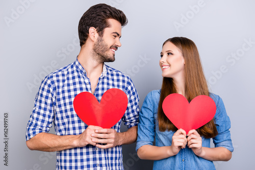 Love story. Trust and feelings, emotions and joy. Happy young couple in love is posing, wearing casual shirts, holding big red papers hearts and smiling on the pure background