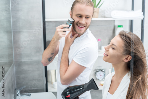 Handsome young man shaving beard with electric razor and smiling woman drying hair with hair dryer