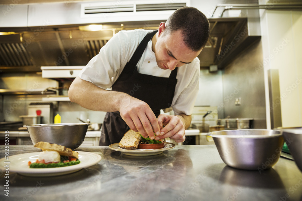 Chef standing in kitchen, wearing apron, plating food. Chef standing in kitchen, wearing apron, plating food.