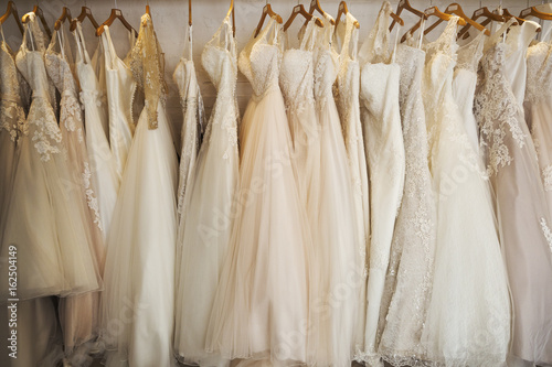 Rows of wedding dresses on display in a specialist wedding dress shop. A variety of colour tones and styles, fashionable lace and boned bodices.  photo