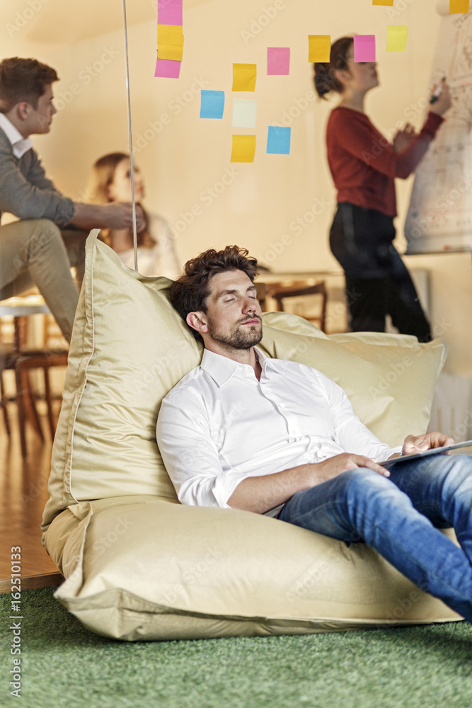 Relaxed man in office sitting in bean bag with meeting in background