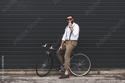 stylish man in sunglasses smoking cigarette and sitting on bicycle