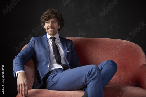 Studio shot of a confident young businessman wearing suit while sitting on sofa at black background.