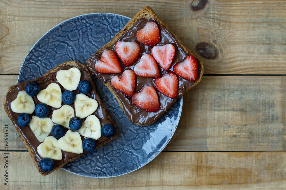 Sweet toasts with heart shaped strawberry and banana, blueberry, chocolate cream in a small plate on wooden table. Breakfast sandwiches Sweet toasts with heart shaped strawberry and banana, blueberry, chocolate cream in a small plate on wooden table. Breakfast sandwiches
