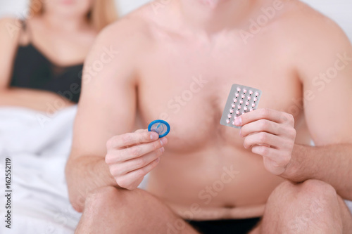 Young man sitting on bed with birth control pills and condom. Concept of choosing the contraceptive method