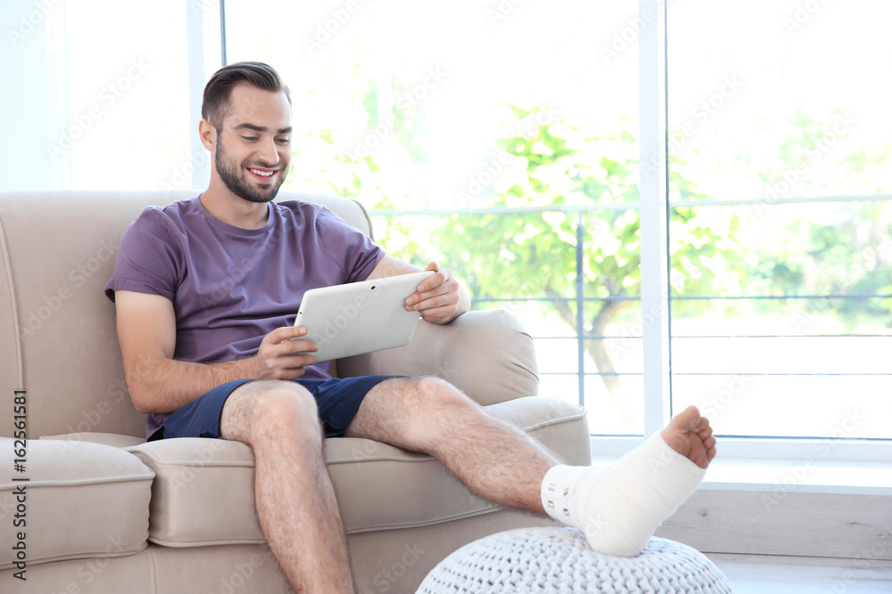 Handsome young man with bandaged leg and tablet computer resting on sofa at home Handsome young man with bandaged leg and tablet computer resting on sofa at home