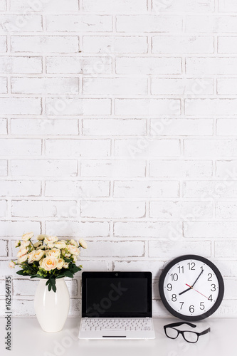 workplace - laptop with blank screen, office clock, glasses and flowers over white wall