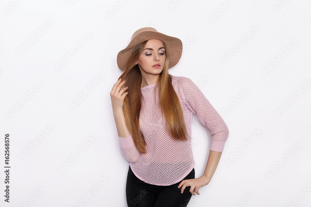 Cute and stylish beautiful young blonde woman in trendy clothing practicing model poses and looking away with smile while standing against white wall background Cute and stylish beautiful young blonde woman in trendy clothing practicing model poses and looking away with smile while standing against white wall background