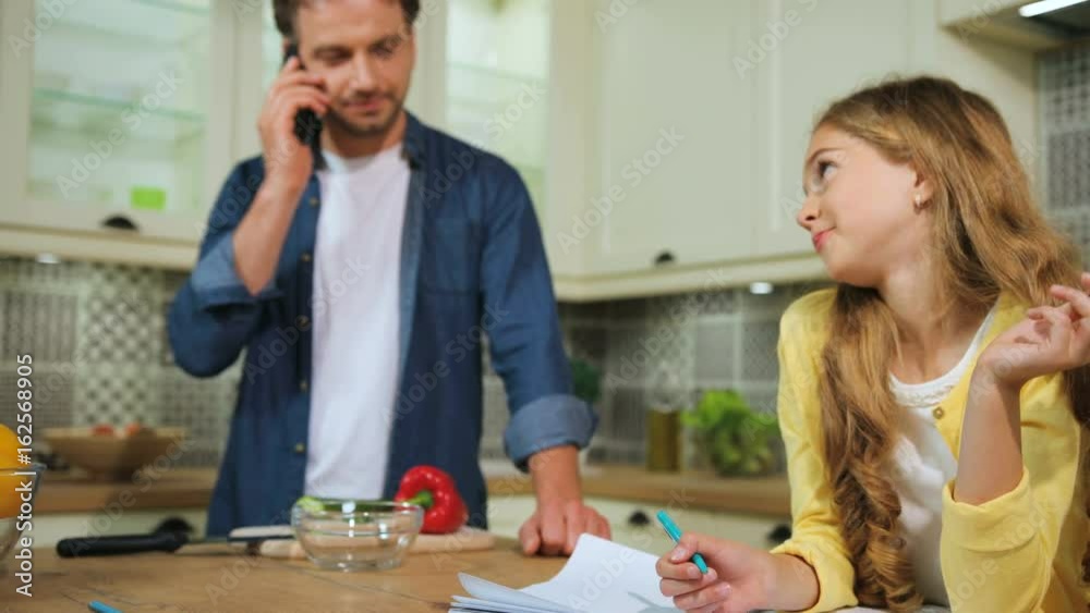 Young girl with blonde hair doing the homework at the kitchen while her father talking on smart phone..