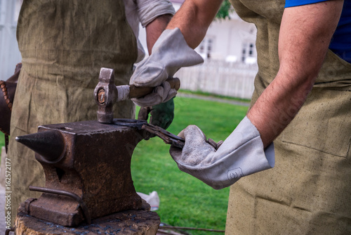 Two blacksmiths near the anvil. One hacks a horseshoe with a hammer. In white gloves and robe. Horizontal frame