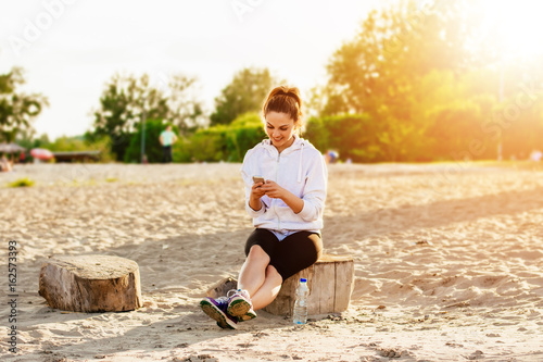 Beautiful girl in sport clothes and using a smartphone and smiling while resting on the beach after workout.