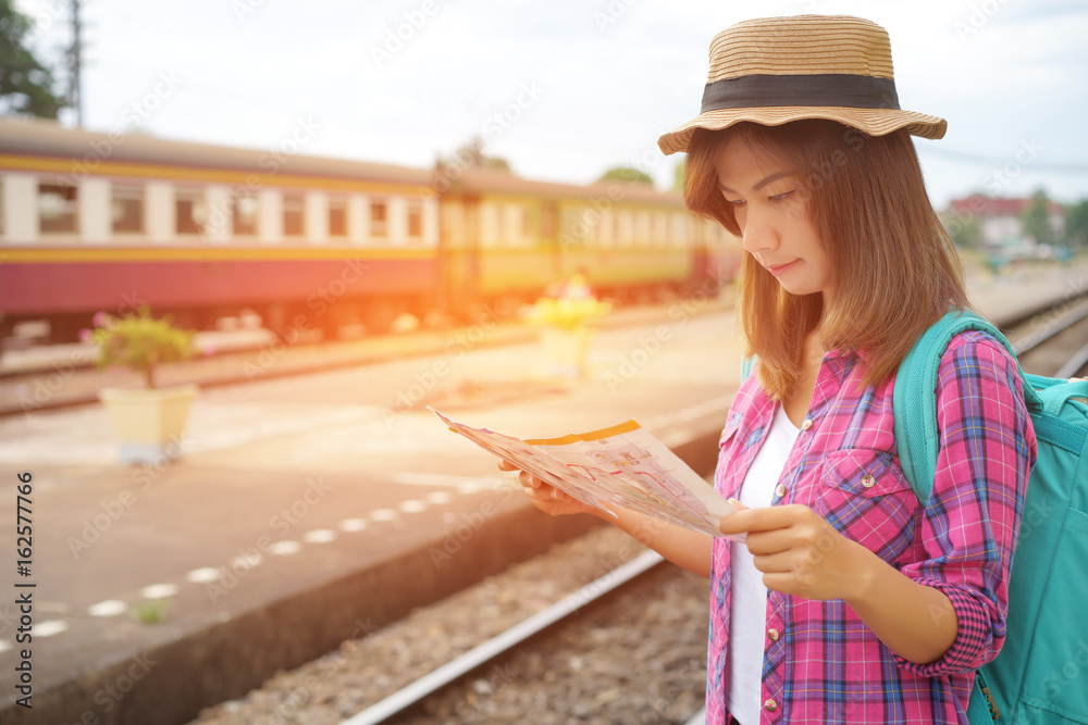 Young woman traveler with backpack and hat at the train station and looking on the map for plan to travel, Travel concept, Sun light flare