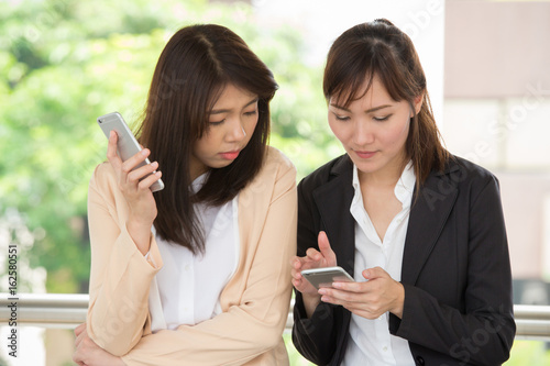 Two business women are discussing about work by using her smartphone to show the information and detail of project. They are standing outside the office.