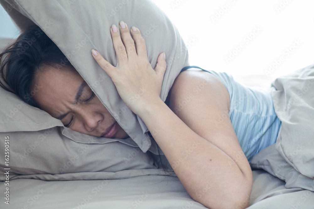Young woman lying in bed suffering from sound covering head and ears with pillow making unpleasant face. early wake up not getting enough sleep - obrazy, fototapety, plakaty 