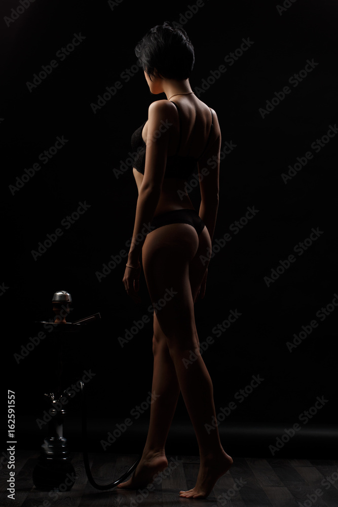 Naklejka premium Sexy, beautiful and attractive woman with gorgeous slim body wearing lacy underwear is posing in the dark studio. Appealing female model with seductive figure and short haircut in the black lingerie