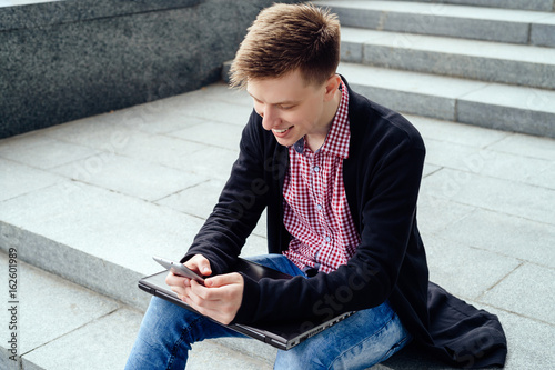 Happy handsome young man sitting on the stairs and using smart phone in the city outdoors.  Student rests after college outdoors © mirage_studio