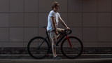 Tattooed biker hipster man in shorts standing against a grey wall next to a fixed gear bike