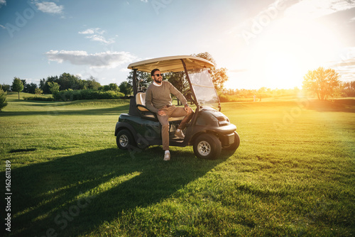 Handsome bearded man in sunglasses sitting in golf car and looking away