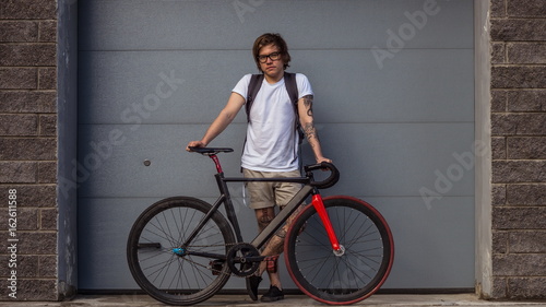 Tattooed biker hipster man in shorts standing next to a fixed gear bike against the background of gray rolling gates