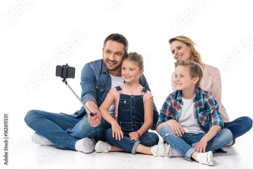 smiling family taking selfie on smartphone while sitting on floor isolated on white