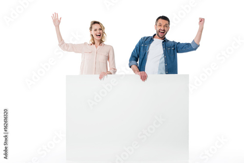 Happy young couple holding blank banner and standing with raised hands isolated on white