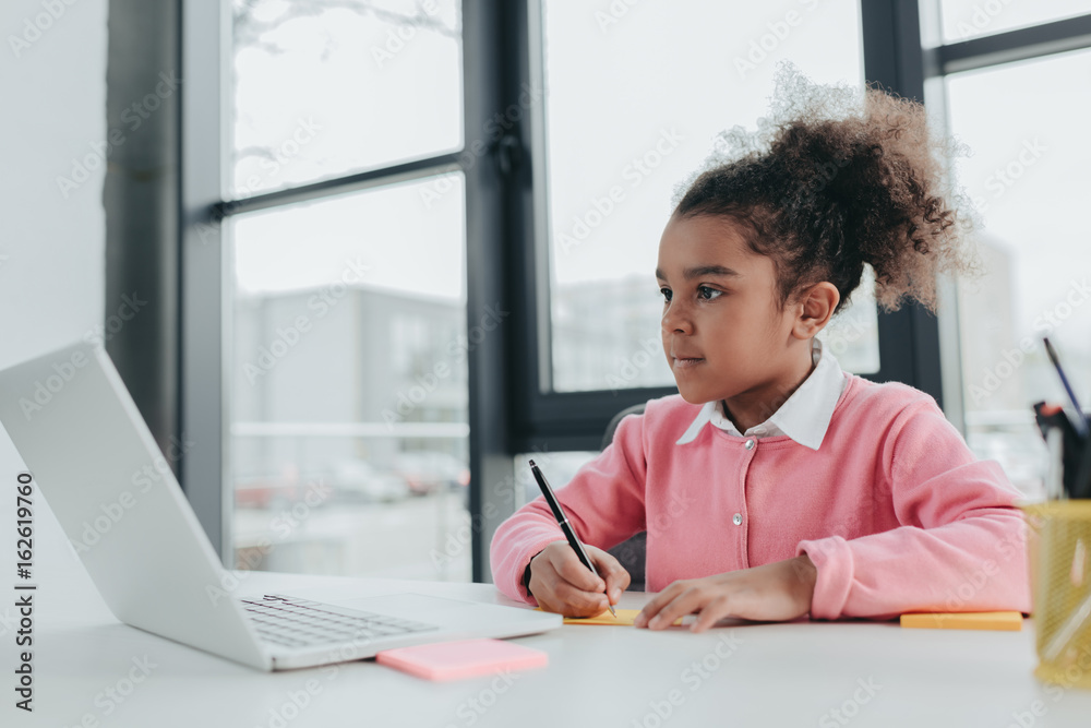 Adorable little african american girl using laptop while sitting at office table Adorable little african american girl using laptop while sitting at office table