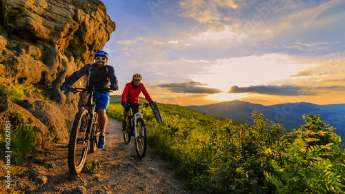 Mountain biking women and man riding on bikes at sunset mountains forest landscape. Couple cycling MTB enduro flow trail track. Outdoor sport activity.