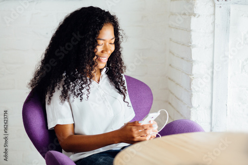 Young gorgeous african american woman with headphones sitting in purple armchair smiling and lookink at smart in modern loft interior.