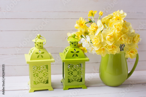 Bright yellow spring daffodils or narcissus flowers in green pitcher and decorative green lanterns on white wooden background.