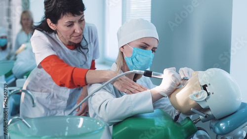 Dentistry student or hygienist practicing on a dummy at a medical school as a qualified teacher demonstrates the technique to her from behind