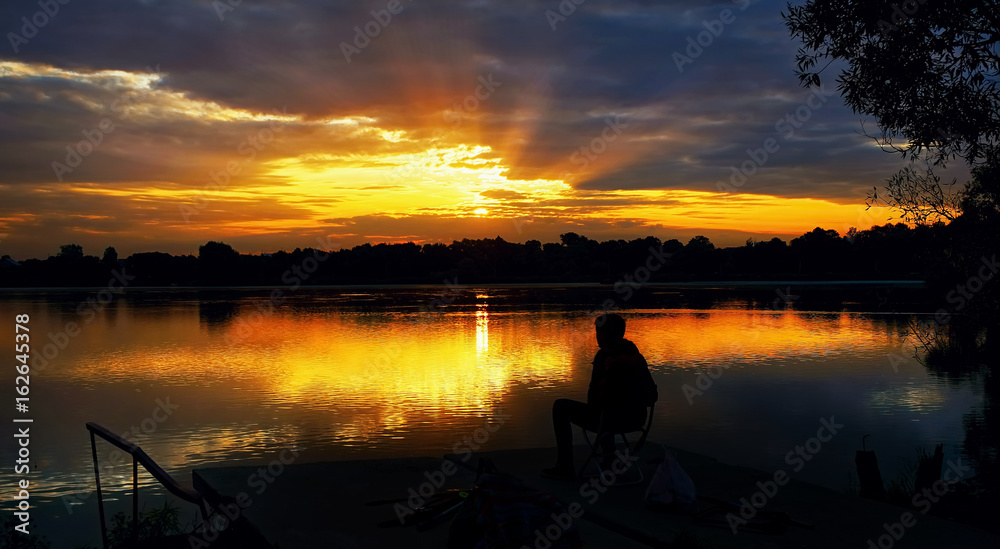 The silhouette of a seated fisherman on a beautiful background of a rising sun with rays in the dramatic clouds .